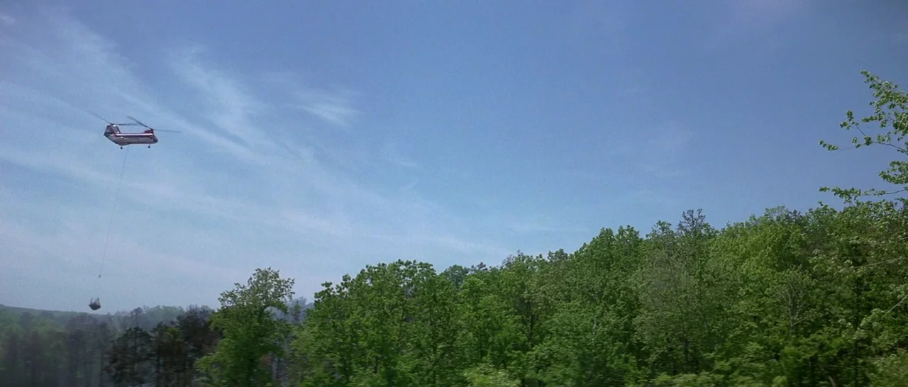 Starman (1984) movie still: wide — A view of a dense deciduous forest stretching under a clear daytime sky. A transport helicopter hovers above the…