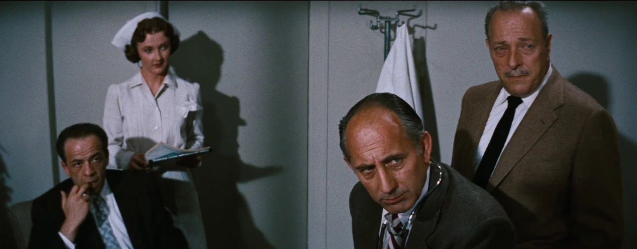Bigger Than Life (1956) movie still: medium — A sterile medical setting features light-colored walls and a wall-mounted garment hook. Four people…