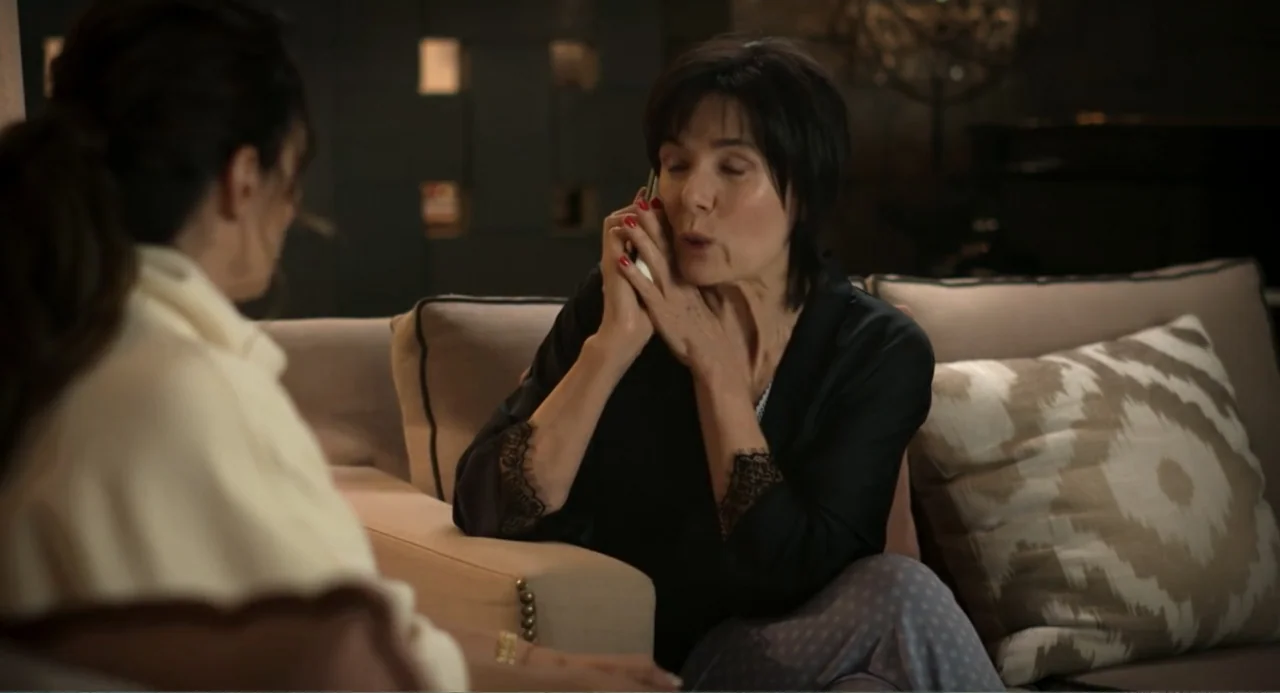La posada (2023) movie still: medium — A dimly lit room features a light-colored sofa with patterned throw pillows. A woman in a black blouse sits…