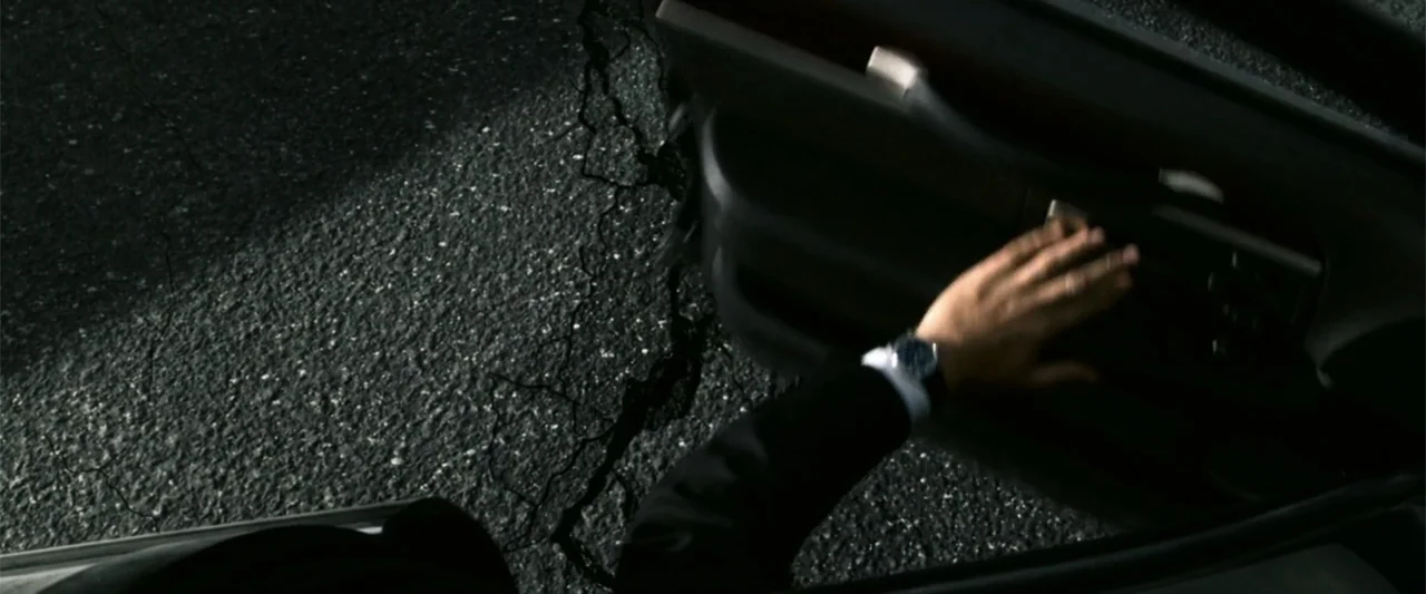 2012 (2009) movie still: closeup — A dark asphalt surface is illuminated by a single harsh light at night. A hand in a suit sleeve reaches to pull a…