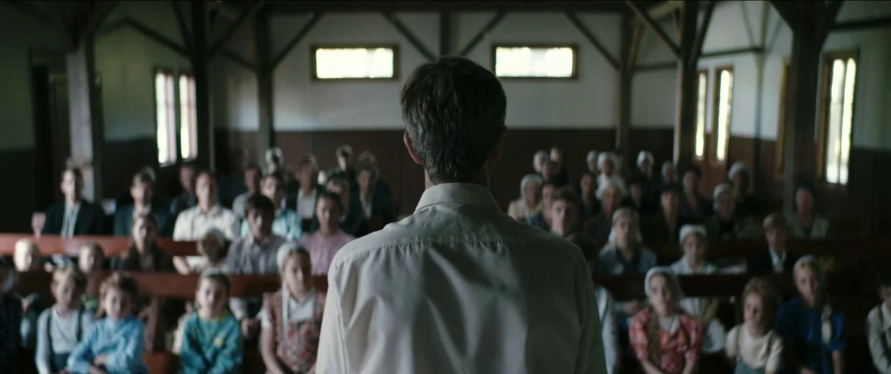 Good Favour (2017) movie still: medium — A wooden interior space with long rows of benches filled with people of varying ages. A man stands in the…