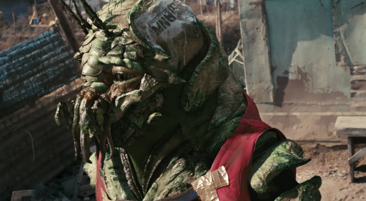 District 9 (2009) movie still: closeup — A grimy alien character with green scales stands in a cluttered, impoverished outdoor setting. The creature…