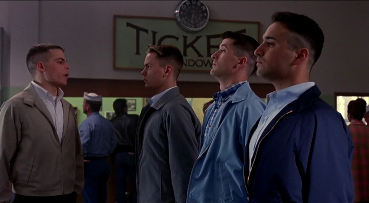 Dogfight (1991) movie still: medium — An interior shot of a 1950s public transit station with a ticket window. Four young men in matching-style…