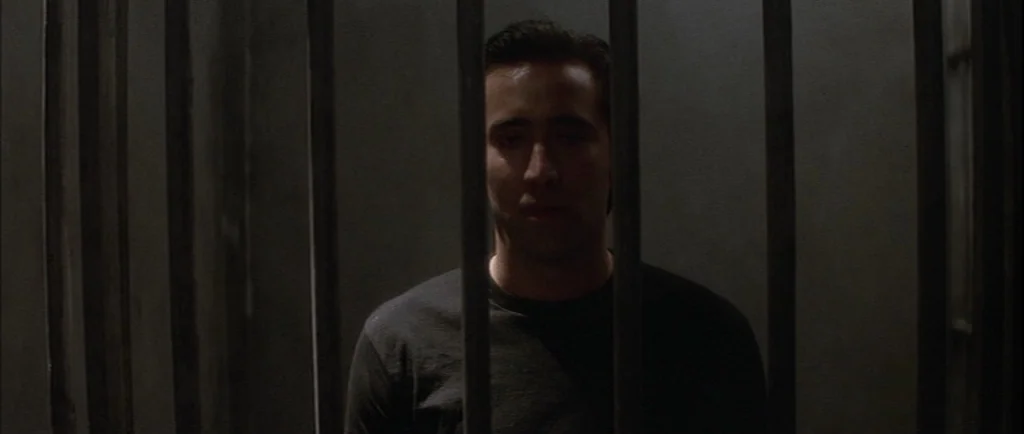 Wild At Heart (1990) movie still: medium — A dark space defined by vertical metal bars in the foreground. A man stands behind the metal bars looking…