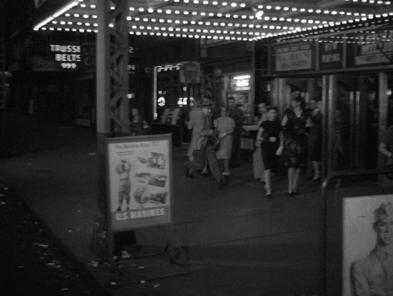 The Naked City (1948) movie still: wide — A 1940s theater entrance at night with illuminated marquee lights and a poster stand. Several people walk…