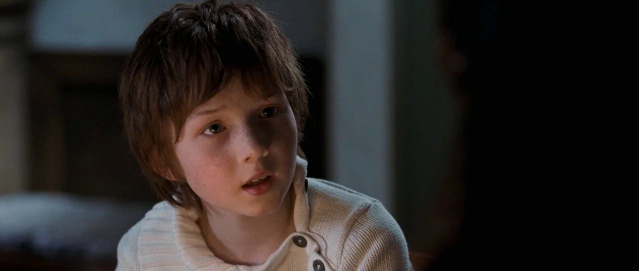 Womb (2010) movie still: closeup — A dimly lit indoor space with shadows obscuring parts of the background. A young child with freckles and brown…