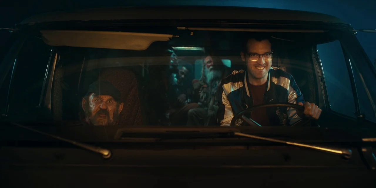 Destroy All Neighbours (2024) movie still: medium — The front cabin of a car during the night with dark, shadowy surroundings. A man in glasses…