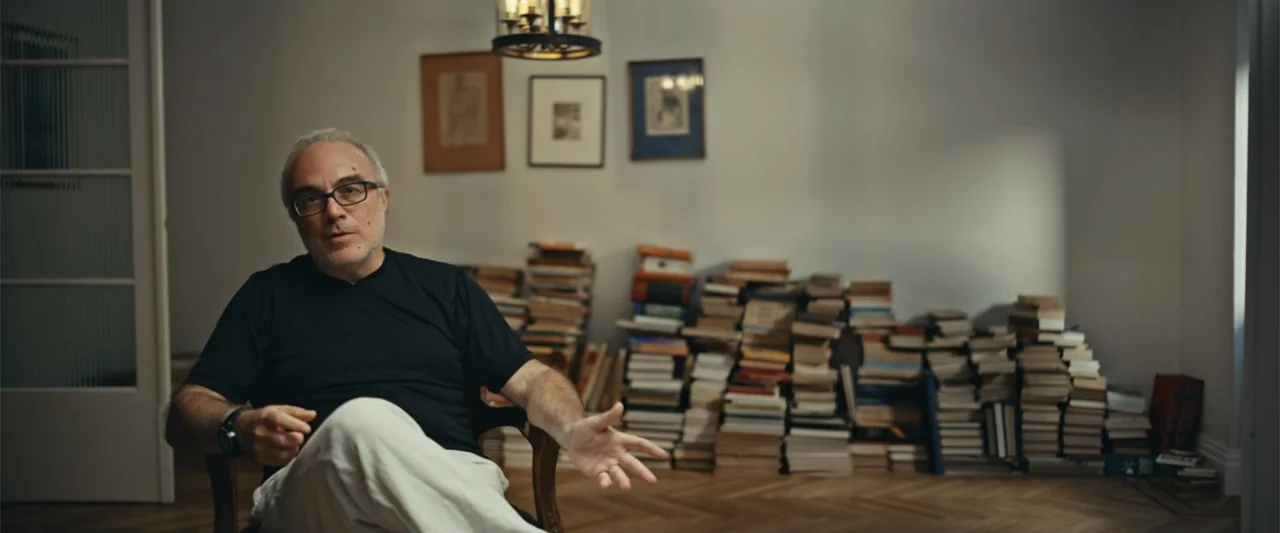 Giannis: The Marvelous Journey (2024) movie still: medium — An interior space contains a man seated in a wooden chair surrounded by stacks of books…