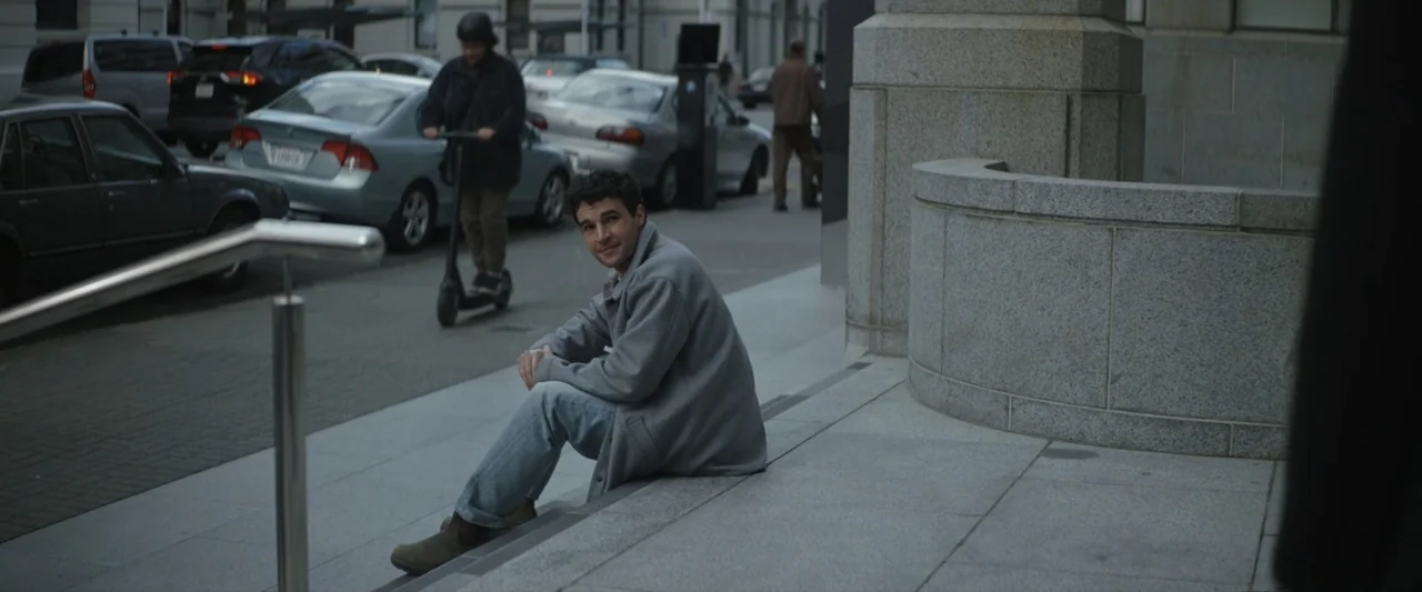 Wolf Man (2025) movie still: medium — A man sits on stone steps outside a gray urban building with cars nearby. He turns to look at the camera while…