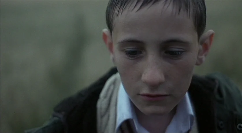 Ratcatcher (1999) movie still: closeup — An open grassy field under an overcast, diffused sky. A young boy looks down toward the ground with a…