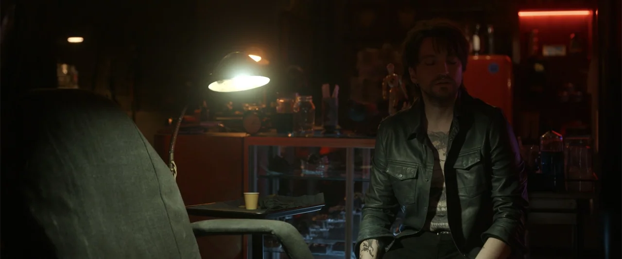Final Destination Bloodlines (2025) movie still: medium — A dimly lit tattoo studio features a desk lamp and a glass display cabinet. A young man…
