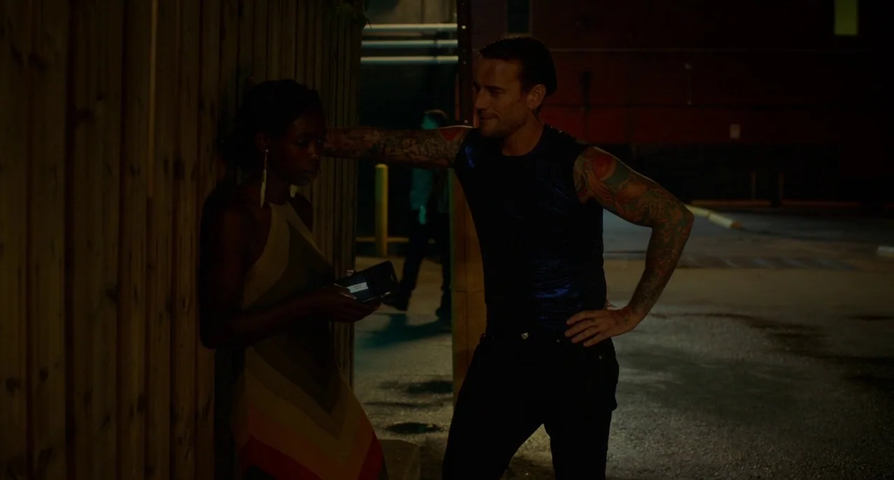 Rabid (2019) movie still: medium — A dark outdoor parking lot at night beside a vertical wooden privacy fence. A man in a blue sleeveless shirt…