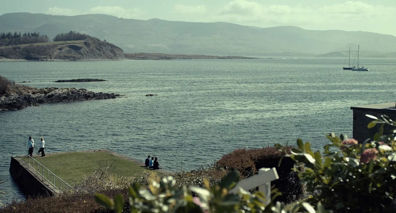 The Lobster (2015) movie still: wide — A grassy concrete pier extends into a calm bay surrounded by rolling coastal hills. Five people stand or sit…