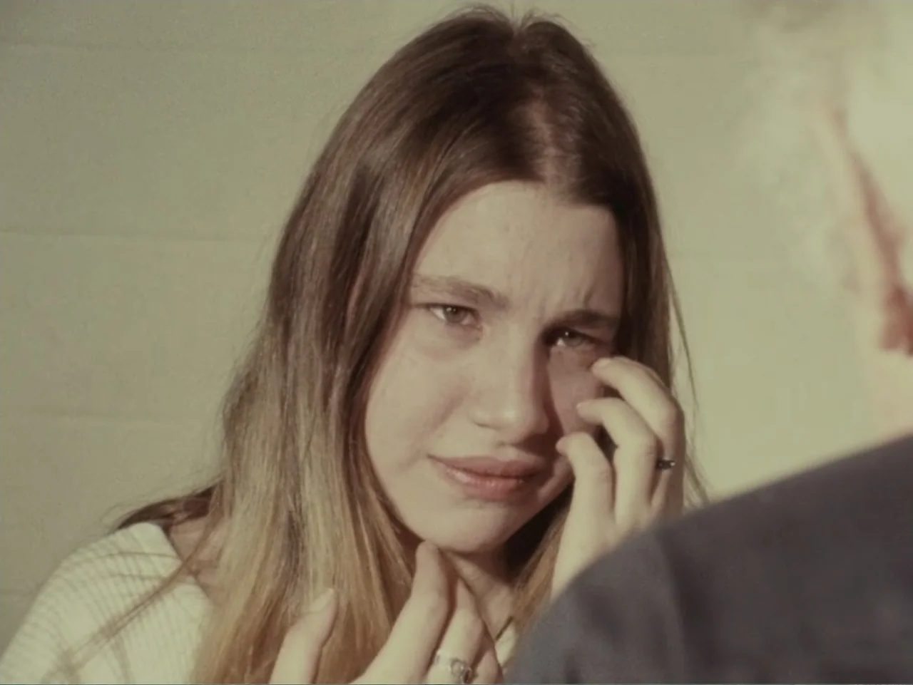 Don't Expect Too Much (2011) movie still: closeup — A plain interior room with a textured white wall in the background. A young woman with long hair…