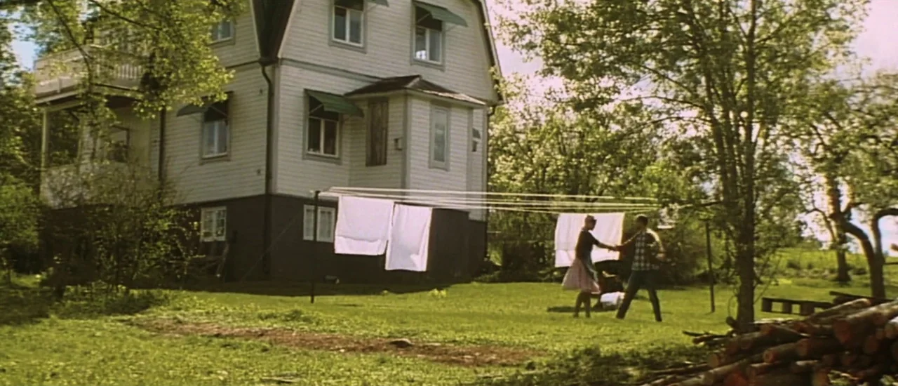 Dancer in the Dark (2000) movie still: wide — A white two-story wooden house stands in a grassy yard near a forest. A man and woman walk together…