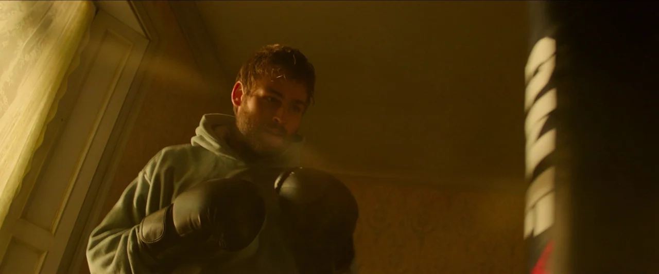 Unwelcome (2022) movie still: medium — A dimly lit room contains patterned wallpaper and a suspended punching bag. A man with boxing gloves stands…