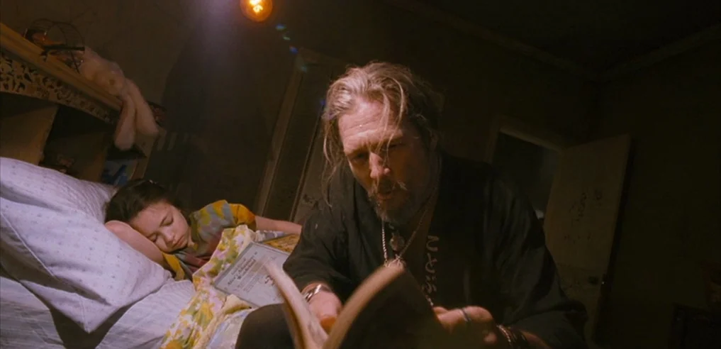 Tideland (2005) movie still: medium — A dark, dimly lit space with a bed, pillows, and a hanging light fixture. An older man sits by a child's…