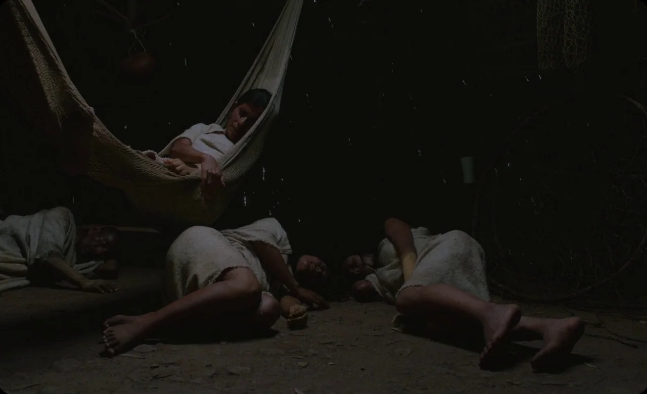 Eureka (2024) movie still: wide — A dark, dimly lit earthen floor structure contains four people sleeping on the ground. One person rests in a woven…