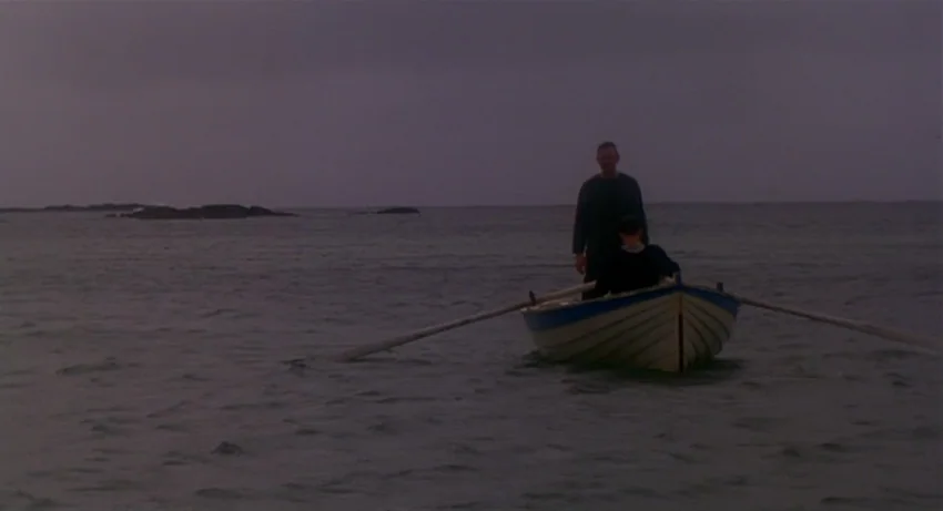 The Secret Of Roan Inish (1994) movie still: wide — A wooden rowboat sits in calm, dark water under an overcast sky. A man stands in the boat while…