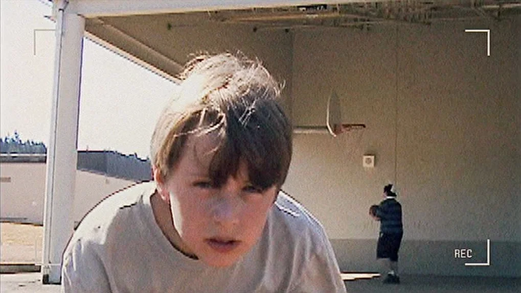 Mean Creek (2004) movie still: closeup — An outdoor paved area beside a beige building features a basketball hoop mounted overhead. A teenager…