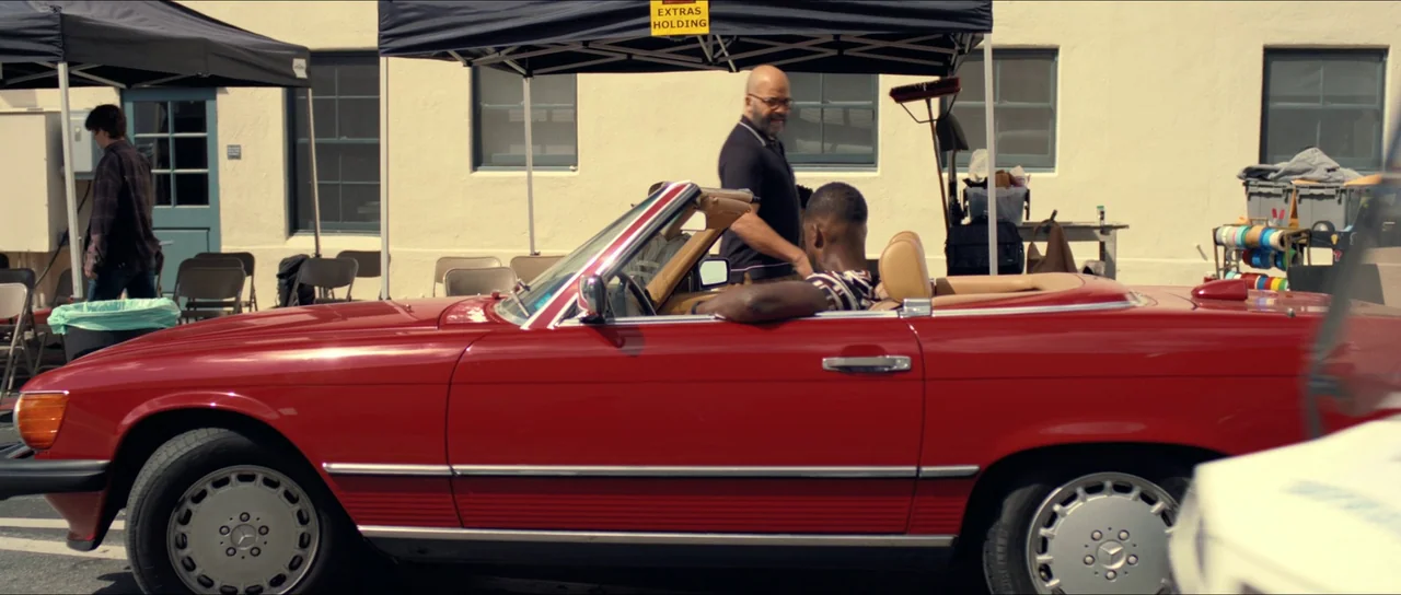 American Fiction (2023) movie still: wide — An outdoor parking area features a red convertible parked beneath a black pop-up tent. Two men interact…