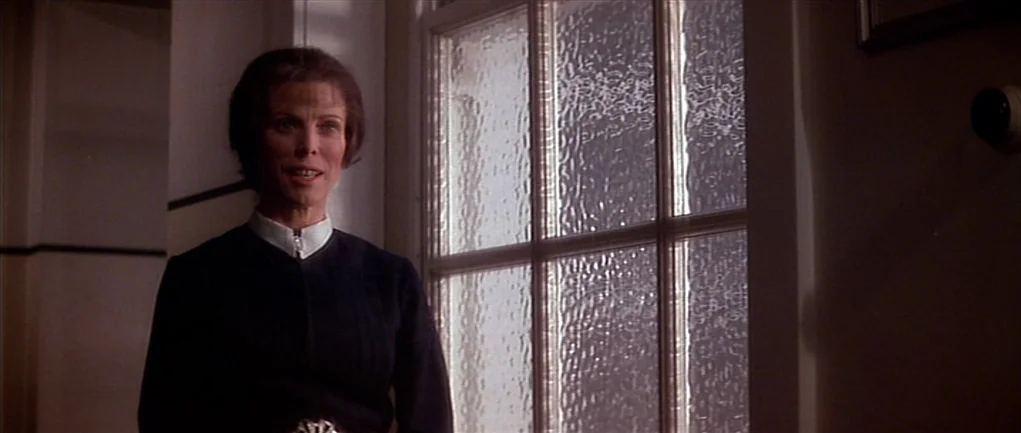 The Omen (1976) movie still: medium — An interior hallway features white walls and a large paneled window with frosted glass. A woman stands in the…