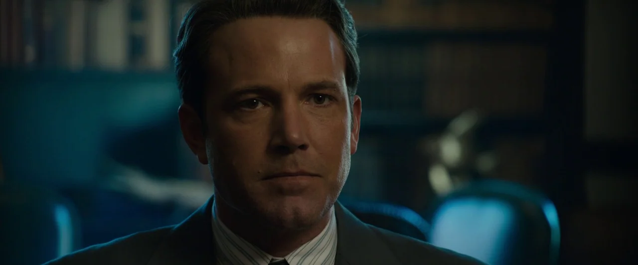 Live By Night (2016) movie still: closeup — A dimly lit room with rows of dark wooden bookshelves in the blurred background. A man sits at a desk…