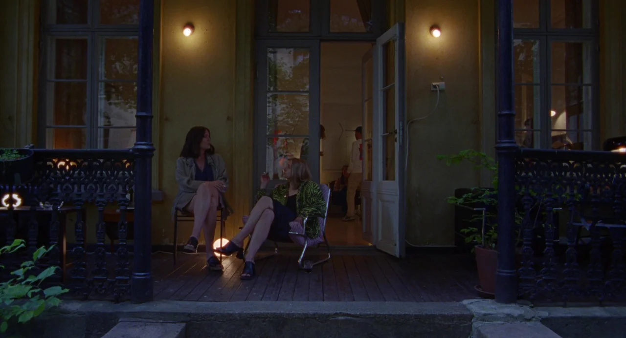 Sick of Myself (2022) movie still: wide — A wooden porch features two women sitting on chairs in front of glass doors. The interior space behind the…