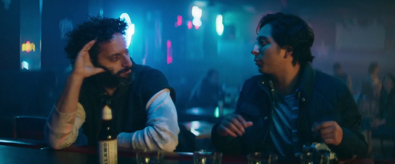 The Long Dumb Road (2018) movie still: medium — A dimly lit bar space filled with neon signs and blurred background figures. Two men sit at the…