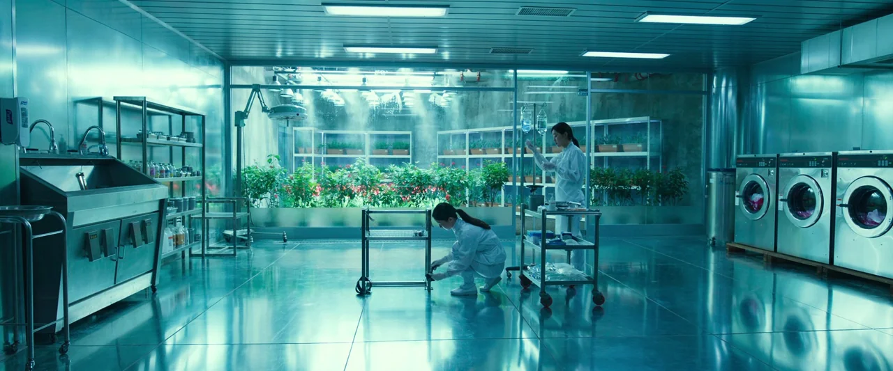 Believer (2018) movie still: wide — A clinical laboratory space features a glass-walled greenhouse partition containing rows of plants. Two…