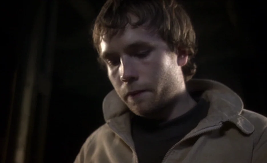 Dear Wendy (2005) movie still: closeup — A dark, shadowed space with wooden rafters visible in the background. A young man looks downward with a…
