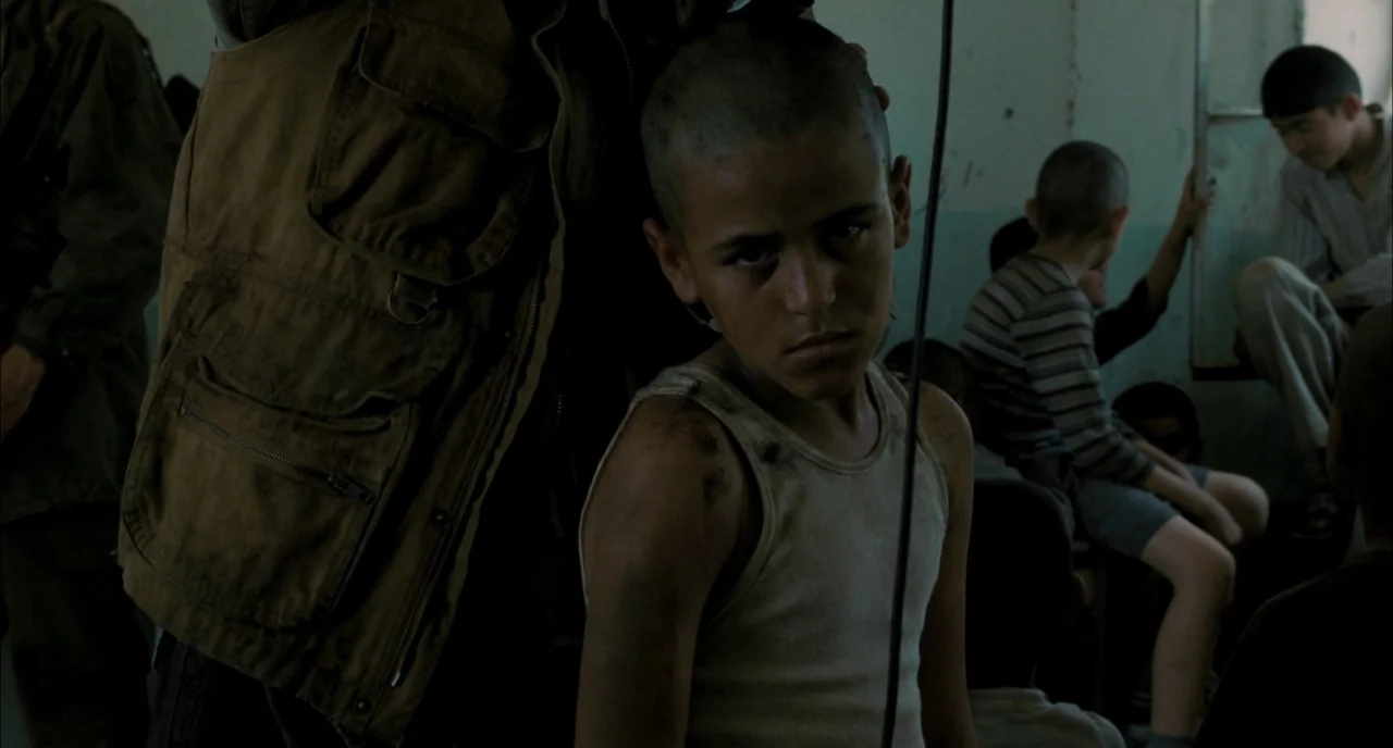 Incendies movie still: medium — A crowded, dimly lit indoor space with several children present in the background. A young boy in a dirty white tank…