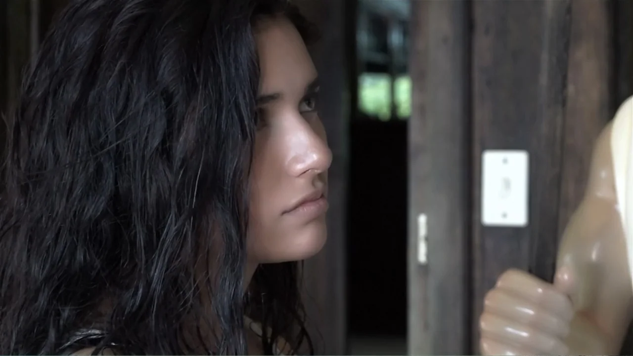 El Duende Sátiro (2016) movie still: closeup — An interior corridor with dark wooden walls and a visible light switch plate. A young woman stands…
