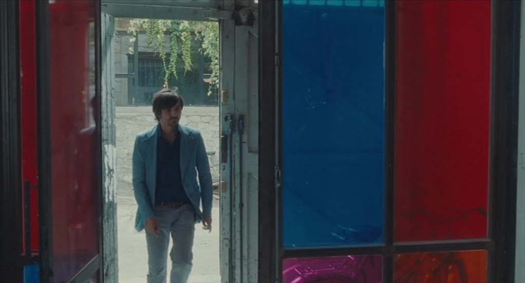 Something in the Air (2012) movie still: medium — A man walks through a doorway featuring panes of red and blue tinted glass. The exterior path…