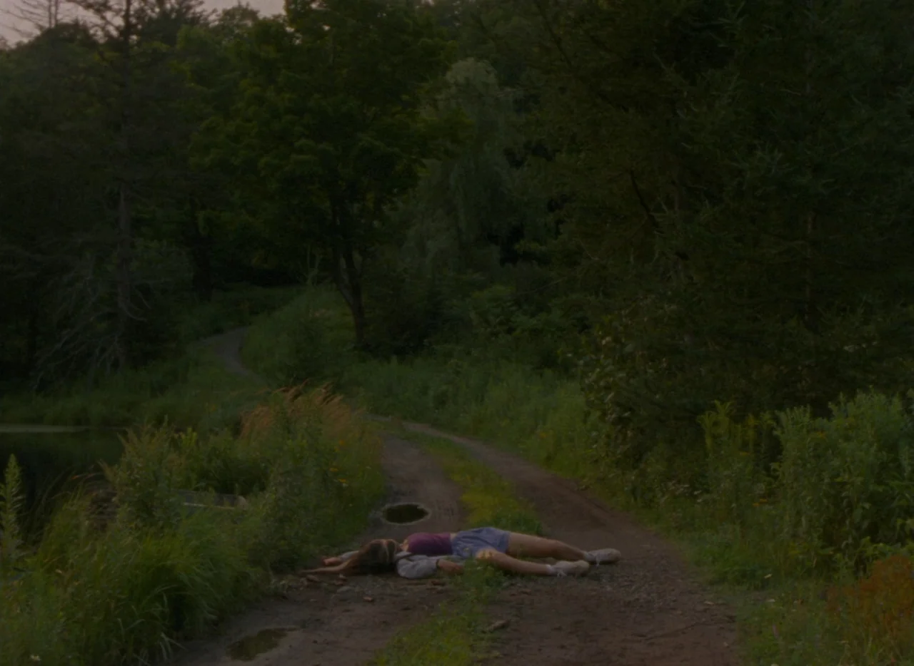 Falcon Lake (2022) movie still: wide — A rural dirt track is flanked by dense woods and tall wild grasses. A person lies flat on their back in the…