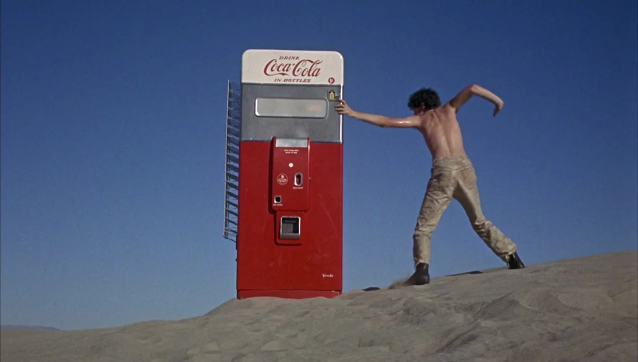 Head (1968) movie still: wide — A vintage Coca-Cola vending machine sits half-buried in a vast, empty sandy desert. A shirtless man stands beside…