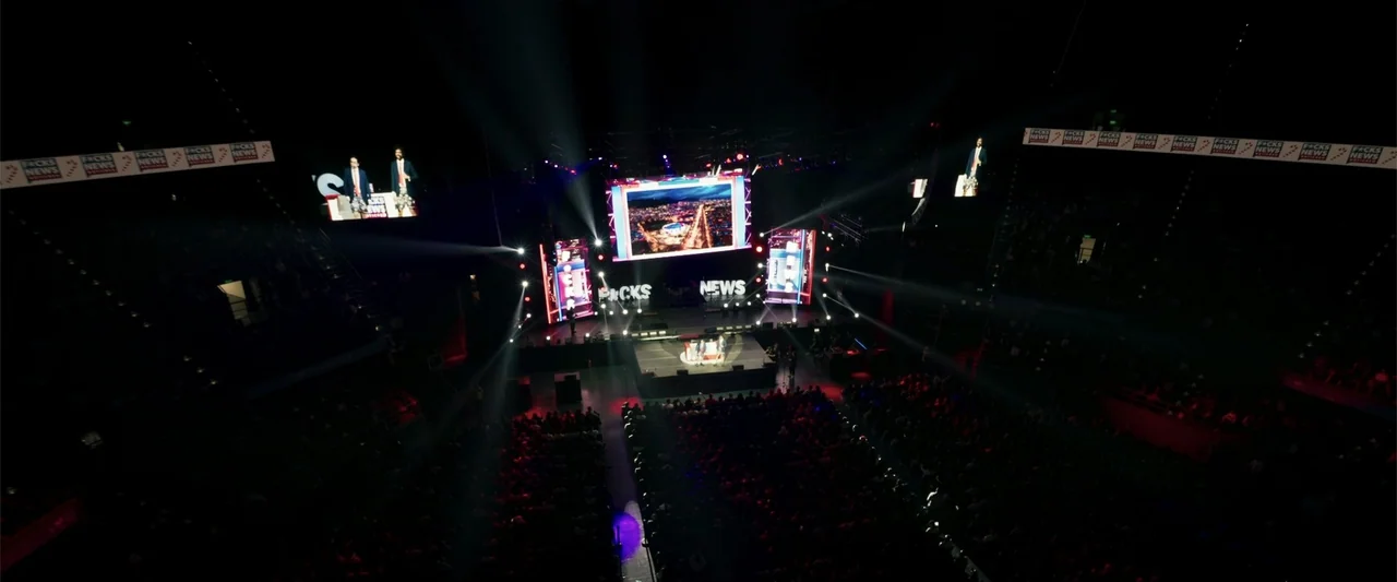Making of: F*CKS NEWS (2025) movie still: wide — A large indoor arena is darkened with a illuminated central stage and audience seating. Multiple…