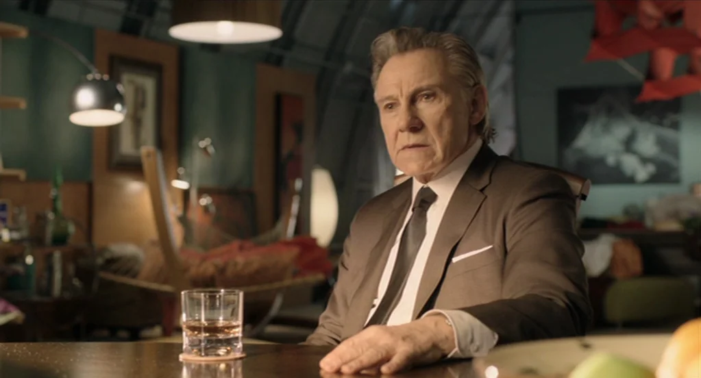 The Congress (2013) movie still: medium — An older man sits at a table in a dimly lit, mid-century style room. He rests his hand near a glass of…