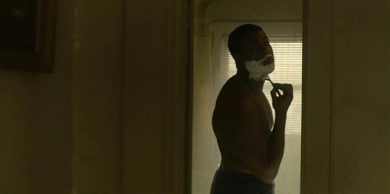 They Cloned Tyrone (2023) movie still: medium — A man stands in a dark room illuminated by light filtering through window blinds. He faces the…