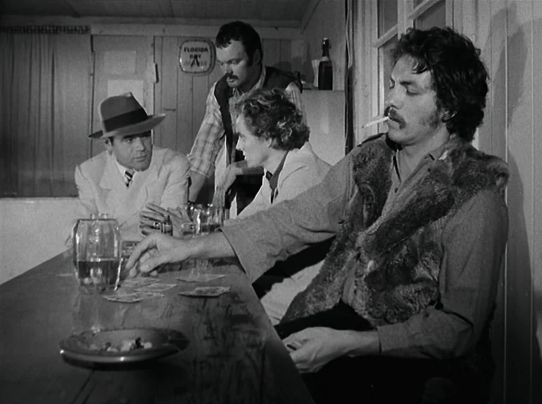 The American Soldier (1970) movie still: medium — A dimly lit room with wood-paneled walls and a wooden bar surface. Four men gather around the bar…