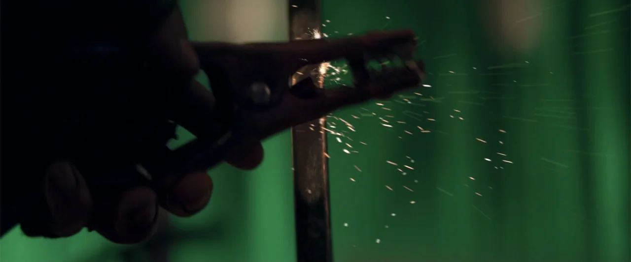 Human Animals (2020) movie still: closeup — A close view shows a metallic terminal clamped against a vertical rod. Bright white sparks shower…