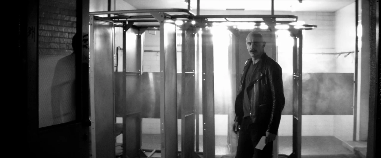 Zeroville (2019) movie still: medium — A communal washroom features metallic shower stalls with bright light filtering through gaps. A man in a…
