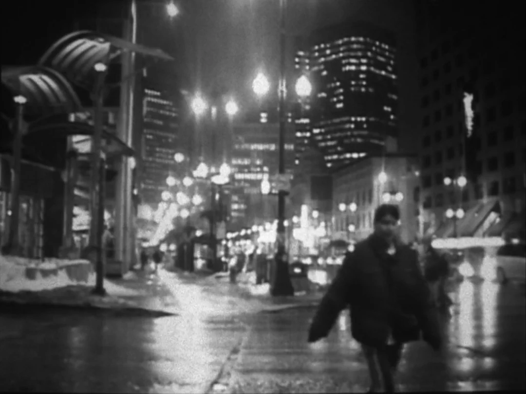 My Winnipeg (2007) movie still: medium — An urban city street at night with wet pavement and tall buildings in distance. A person in a puffy jacket…