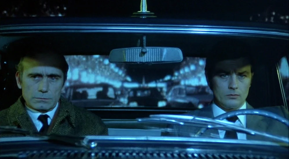 Un Flic (1972) movie still: medium — Two men occupy the front seats of a vehicle driving at night. Blue streetlamp light reflects against the…