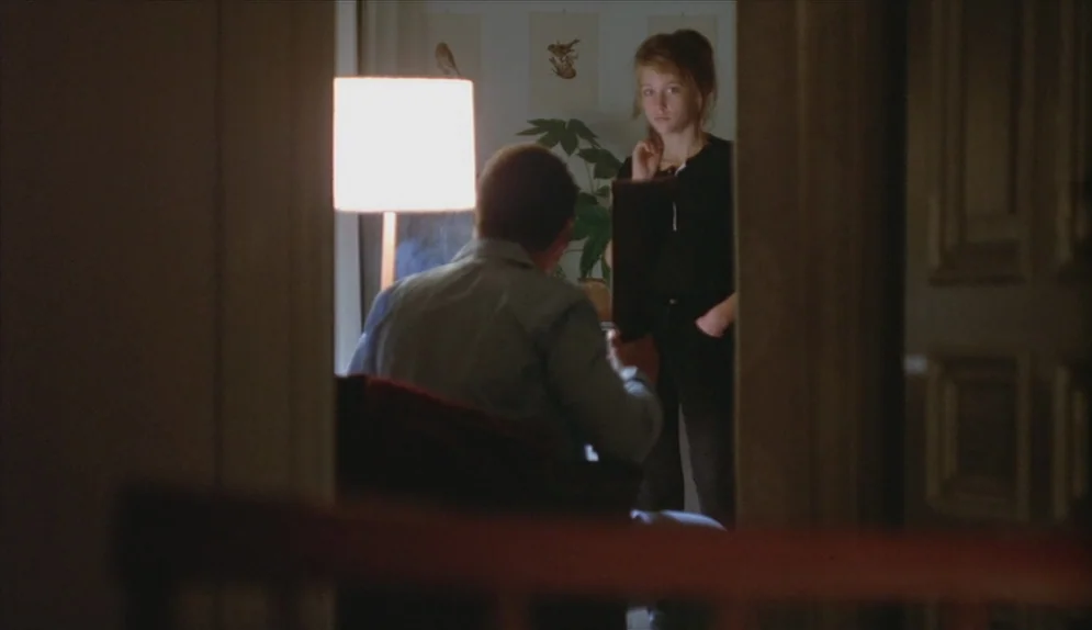 A Swedish Love Story (1970) movie still: medium — A dimly lit room viewed through a dark wooden door frame. A man sits in a chair while a young…