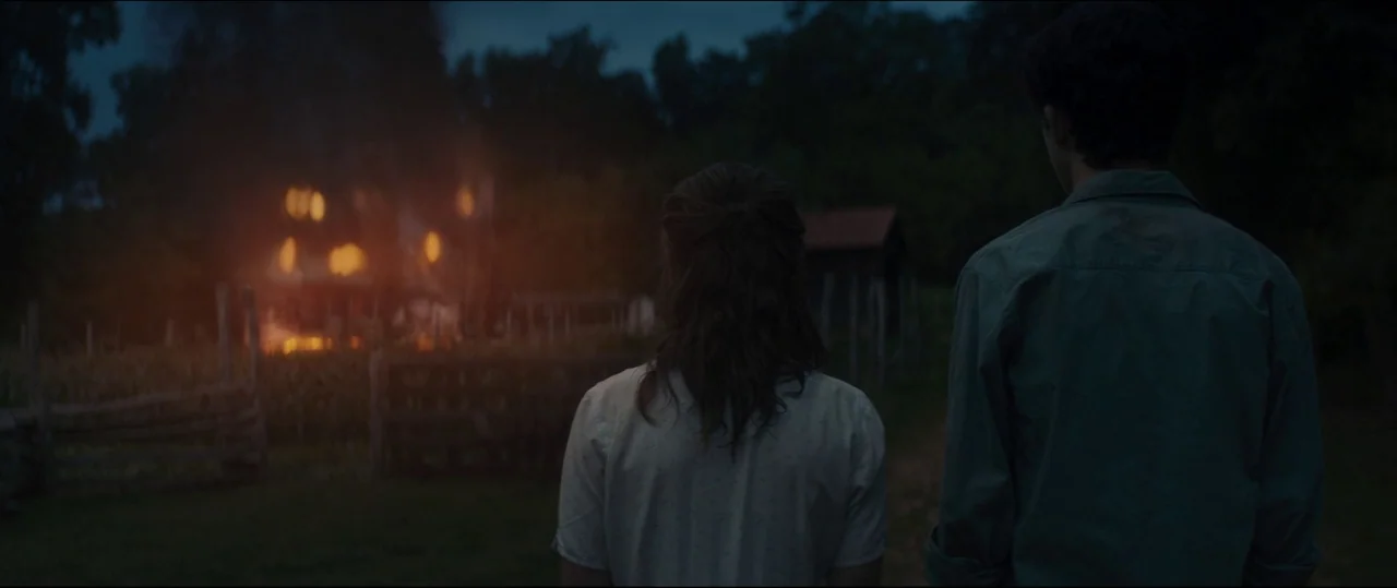 One & Two (2015) movie still: medium — Two individuals stand in a dark field at night, facing a distant burning structure. A wooden fence separates…