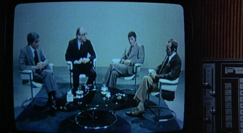 The Passenger (1975) movie still: wide — A 1970s television talk show set features four men seated on minimalist chairs. They are engaged in a…