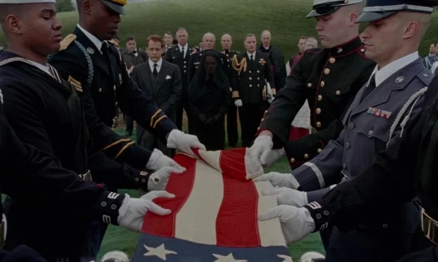 Jackie (2016) movie still: medium — A group of military personnel in formal dress uniforms stand in a cemetery. They hold a folded American flag…