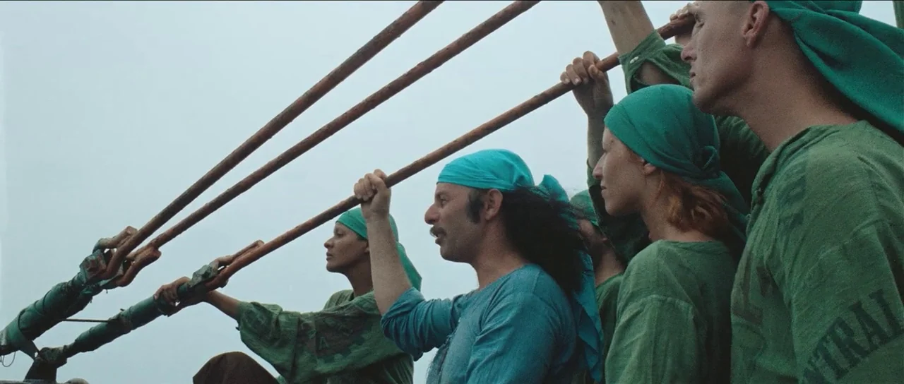 The Holy Mountain (1973) movie still: medium — A group of people stand on an outdoor deck against a pale overcast sky. Several individuals in green…