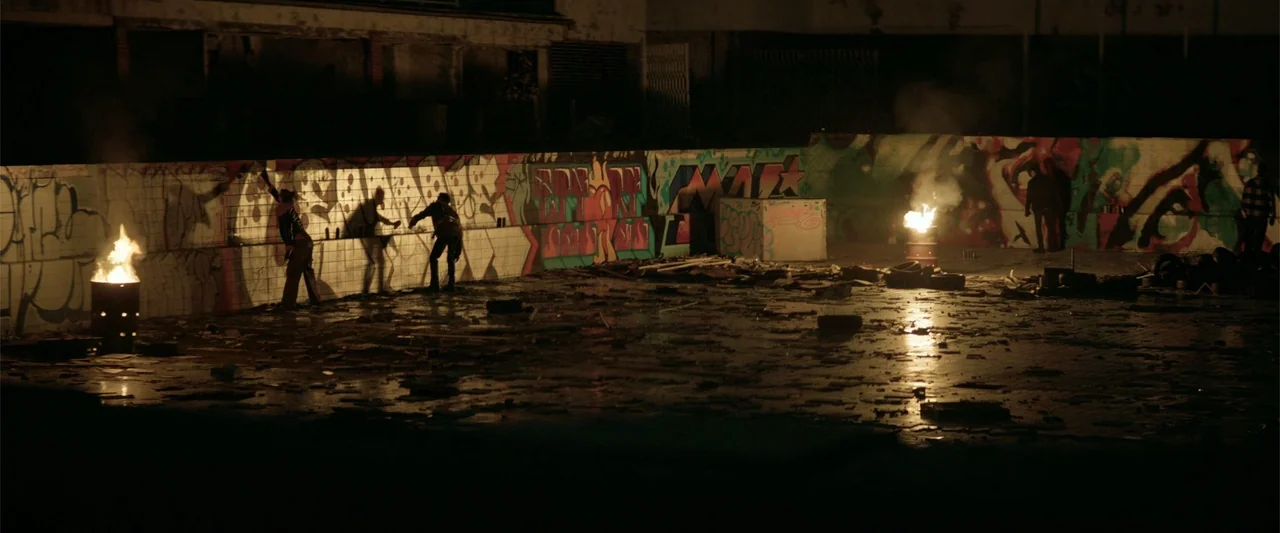 Marked Men: Rule + Shaw (2025) movie still: wide — An abandoned urban lot features a long graffiti-covered wall lit by two fire barrels. Several…